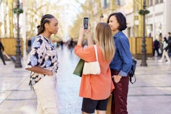 Young multiracial content female friends in stylish clothes taking self portrait on phone while standing on street with paper bags. Diverse women taking selfie on smartphone in street