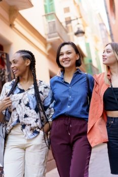 Positive multiracial female friends in elegant outfit smiling during stroll on blurred city street. Cheerful diverse ladies spending time in city