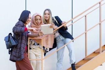Confident young multiracial women smiling and sharing tablet standing on metal staircase on street. Smiling young diverse ladies using tablet on stairs