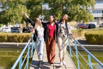 Full body of cheerful multiracial female friends strolling together on wooden footbridge over river in city on sunny summer day. Positive diverse women walking on footbridge