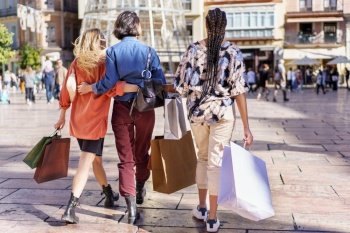 Back view of unrecognizable trendy young female friends in stylish outfits cuddling while walking on city square with paper bags after successful shopping. Anonymous stylish ladies walking on square with shopping bags