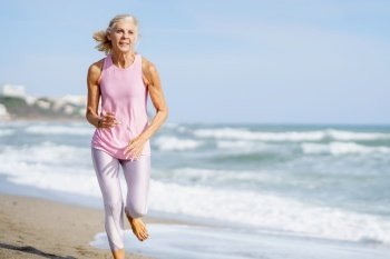 Content senior female in sportswear running on sandy seashore and training on sunny day in summer. Elderly sportswoman running along seashore in summer