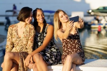 Positive young multiracial female friends in dresses taking self portrait while sitting together near river with boats on summer day. Content diverse girlfriends taking selfie on pier