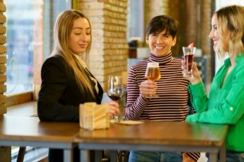 Merry adult female in casual clothes drinking beer and looking at camera with smile while spending time with girlfriends at table near window in pub. Adult woman drinking alcohol with friends in bar