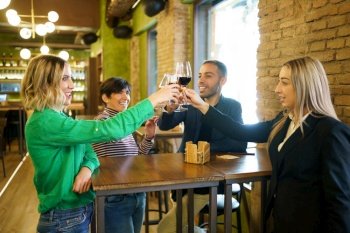 Multiracial man and women smiling and clinking glasses of alcohol beverages over table while proposing toast during meeting in bar. Diverse friends clinking glasses over table in beautiful pub