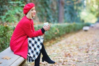 Side view of dreamy female with closed eyes wearing red beret and coat sitting on bench in park with fallen dry leaves and drinking coffee. Stylish woman with takeaway beverage in autumnal park