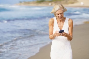Mature female walking on the beach using a smartphone. Elderly woman enjoying her retirement at a seaside location.. Mature female walking on the beach using a smartphone.