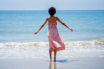 Rear view of black woman with afro hair in summer dress, opening her arms looking out to sea. Girl enjoying her holiday in a coastal area.. Rear view of black woman with afro hair in summer dress, opening her arms looking out to sea.