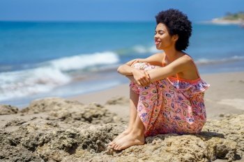 Happy black female sitting on the rocks on the beach looking at the sea smiling. Girl enjoying her holiday in a coastal area.. Happy black female sitting on the rocks on the beach looking at the sea smiling