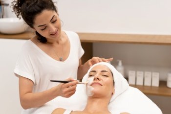 Smiling female cosmetologist applying hydrating mask with brush on face of female customer lying on table in beauty salon. Beautician applying facial mask on client