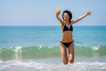 Delighted African American female tourist in swimwear jumping with raised arm while taking self portrait on smartphone near sea in tropical resort. Happy black woman taking selfie while jumping on coast