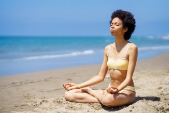 Full body of peaceful slim black female tourist in bikini sitting in Lotus pose on sandy seashore with closed eyes, while meditating during yoga session on sunny summer day. Fit young African American woman in Padmasana meditating on sandy beach
