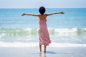 Back view of young woman in stylish summer dress standing on sandy beach with raised arms and looking at rippling ocean. Carefree ethnic female enjoying summer day near sea