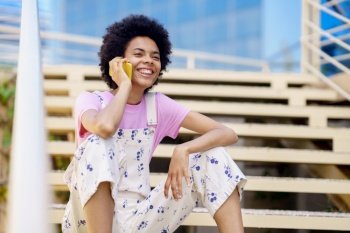 Smiling black woman talking on her smartphone in casual clothes. Happy young female sitting on urban steps.. Smiling black woman talking on her smartphone in casual clothes.