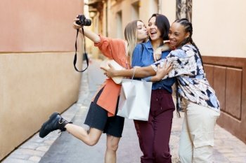 Delightful elegant multiracial women standing against blurred urban building and taking self portrait on camera while rejoicing at purchases. Happy diverse female friends taking selfie on camera on street
