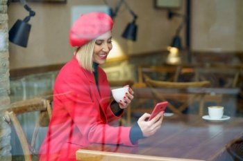 Through glass of fashionable female with cup of coffee text messaging on cellphone while sitting in modern cafe near window. Elegant positive woman with coffee surfing smartphone