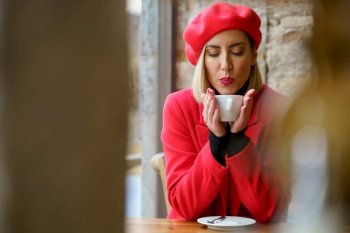 Attractive female in red beret blowing on cup of hot drink in hands while sitting at table in light cafeteria. Woman blowing on hot coffee in cafe