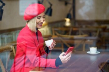 Through glass side vies of smiling female in elegant coat text messaging on cellphone while sitting with cup of coffee at table in cafeteria. Through window of woman browsing smartphone in cafe