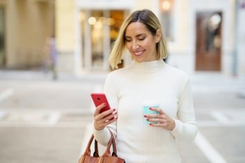Happy female with blond hair wearing white turtleneck browsing cellphone and holding cup of takeaway beverage. Positive female using smartphone and having coffee