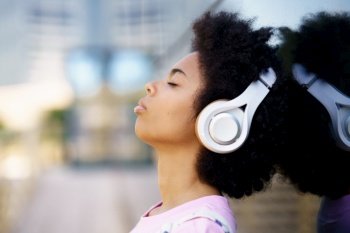 Side view of African American female listening to song in wireless headphones near glass building on street against blurred background. Black woman listening to music on street