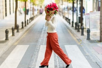 Full body side view of unrecognizable female pedestrian covering face with bouquet of red flowers wrapped in paper while walking on crosswalk on city street. Woman covering face with red flowers while crossing street