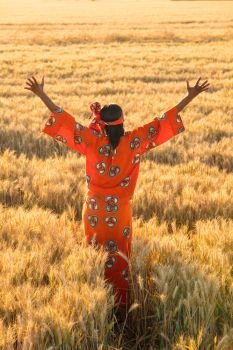 African woman in traditional clothes standing arms raised in field of barley or wheat crops at sunset or sunrise