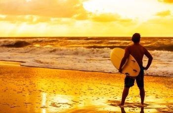 Saturated and stylised rear view of young man male surfer with white surfboard looking at surf on a beach at sunset or sunrise