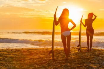 Saturated, stylised, rear view silhouette of two beautiful sexy young women surfer girls in bikini with surfboards on a beach at sunset or sunrise