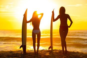 Saturated, stylised, rear view silhouettes of beautiful sexy young women surfer girls in bikinis with surfboards on a beach at sunset or sunrise