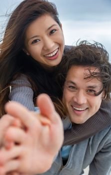Young Asian man and woman romantic couple, boy and girl smiling with perfect teeth playing piggy back on a beach and reaching to the camera