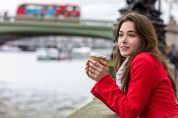 Girl or young woman in a red coat drinking coffee in a disposable cup next to Westminster Bridge with red double decker bus in the background, London, England, Great Britain