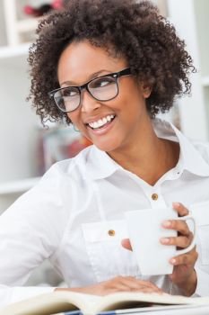Beautiful happy mixed race African American girl or young woman with perfect teeth and wearing glasses, drinking a mug of coffee or tea and reading a book