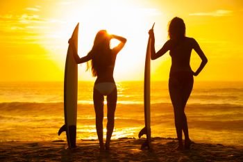 Saturated stylised rear view of two beautiful sexy young woman surfer girls in bikinis with white surfboards on a beach at sunset or sunrise