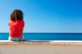 Mixed race African American girl teenager female young woman wearing a red t-shirt and denim shorts sitting on a wall looking out to a blue sea
