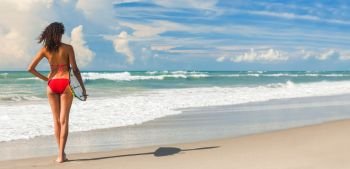 Panoramic banner image rear view of beautiful young woman surfer girl in red bikini with surfboard standing in the surf on a beach 