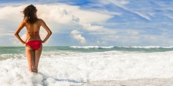 Panorama rear view of a beautiful young woman in bikini standing in the surf waves on a deserted tropical beach with blue sky, panoramic banner image.