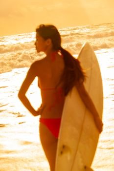 Selective focus rear view of beautiful sexy young woman surfer girl in bikini with white surfboard on a beach at sunset or sunrise the focus is on the waves in the background