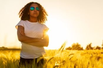 Beautiful mixed race African American female girl teenager young woman wearing sunglassesat sunset or sunrise in a field of wheat, corn or barley