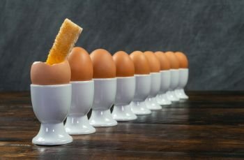 Row of boiled eggs in white egg cups on a wooden table the first egg is broken open with a toast soldier dipped in it