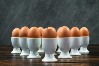 Ten boiled eggs in white egg cups on a wooden table in a triangle arrangement the front egg is broken open