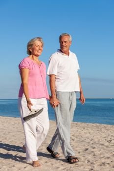 Happy senior man and woman couple walking smiling holding hands on vacation on a deserted tropical beach with bright clear blue sky and calm sea