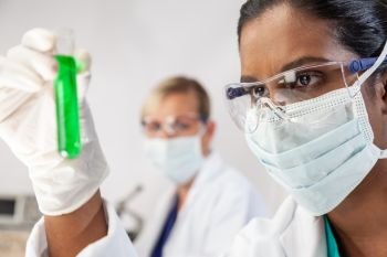 Asian female medical or scientific researcher or doctor looking at a test tube of green solution in a lab or laboratory with her female colleague out of focus behind her.