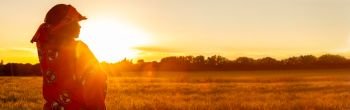 Panoramic web banner African woman in traditional clothes standing, looking, hand to eyes, in field of barley or wheat crops at sunset or sunrise panorama