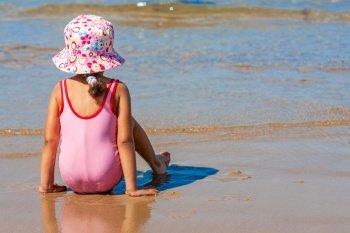 Rear view of cute little girl female child sitting playing on a beach on pink swimming costume and hat