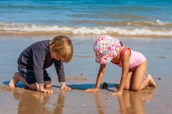 Two cute children little girl female child and boy male child kneeling playing in the sand together on a beach by the seaside