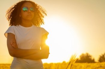 Beautiful mixed race African American female teenager young woman in a field arms folded wearing white t-shirt, sunglasses and back lit by the sun at sunset
