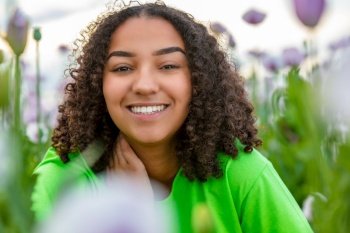 Mixed race beautiful African American biracial girl teenager female young woman wearing green t shirt in field of pink or purple flowers poppies at smiling with perfect white teeth