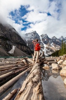 Hiking man with rucsac backpack standing on tree log by Moraine Lake looking at snow covered Rocky Mountain peaks, Banff National Park, Alberta Canada