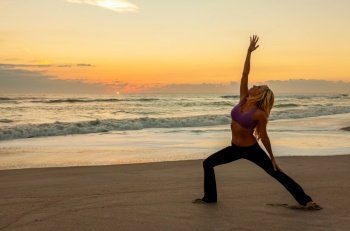 Young woman girl female in a warrior position practicing yoga on a beach at sunrise or sunset
