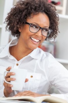 Beautiful happy mixed race African American girl or young woman with perfect teeth and wearing glasses, drinking a mug of coffee or tea and reading a book